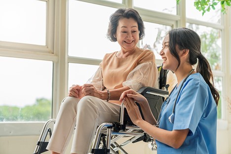 Young woman in scrubs kneels next to older woman in wheelchair