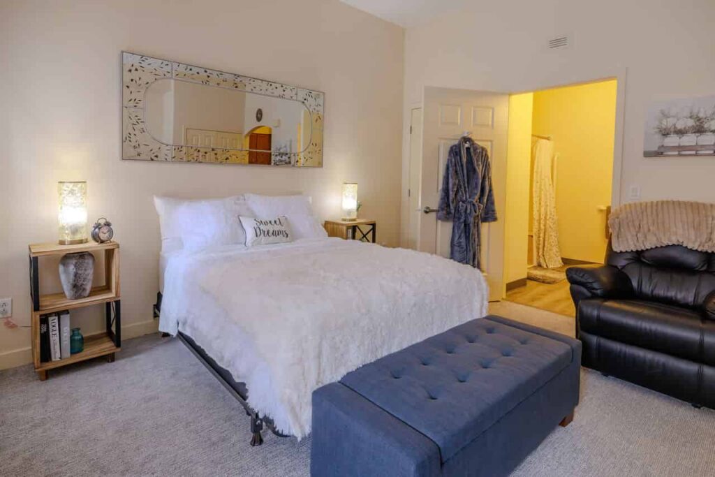 A picture of a bedroom in AlmaVia of Camarillo, an assisted living community. The room has white walls, a beige carpet , a white bed and a personal bathroom.