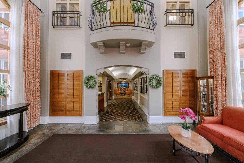 A picture of the lobby of AlmaVia of Camarillo an assisted living community. The room has white walls a balcony, light wood details. On the left a black credenza, on the right a red couch and a coffee table.