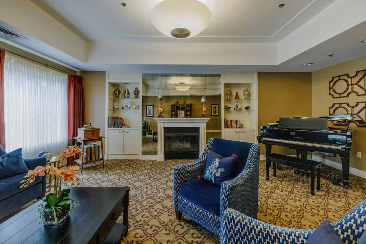 A picture of the common room at AlmaVia of San Francisco, an assisted living community. The room has white walls with tan accents, a tan patterned carpet, a blue living room set, a piano placed in the corner and shelves filled with decor and books.