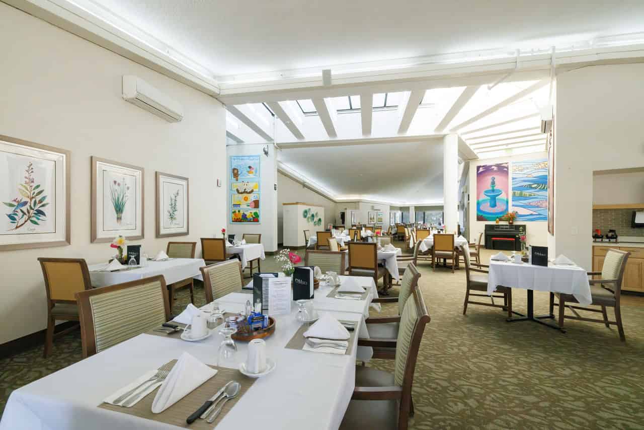 A picture of the dining room at Mercy Retirement and Care Center, an assisted living community. The room has white walls with tan accents, a green patterned carpet, several tables with a white tablecloth and cutlery