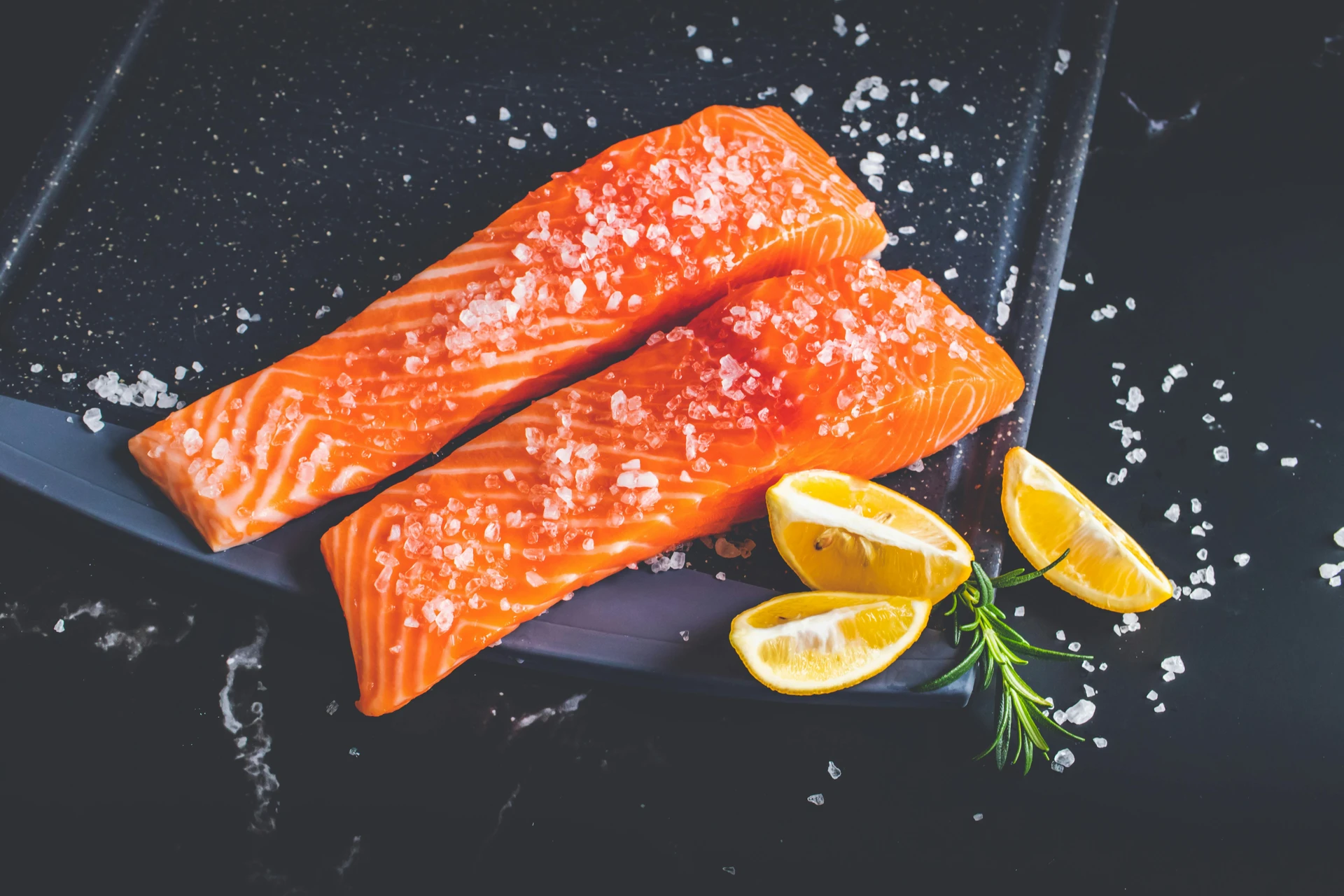 Two raw salmon fillets topped with coarse salt rest on a black tray beside lemon wedges and rosemary—a perfect inspiration for healthy recipes during National Nutrition Month.
