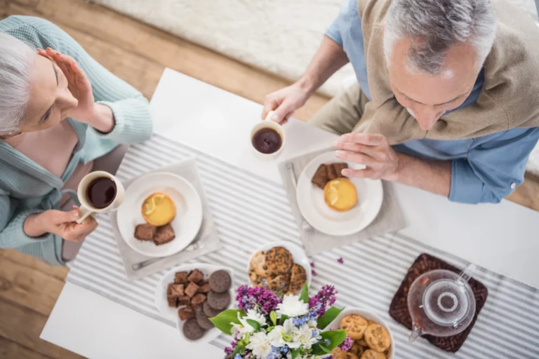 Two older adults sit at a table with coffee, cookies, pastries, and a floral centerpiece on a striped tablecloth, enjoying each other's company while exchanging healthy recipes for National Nutrition Month.