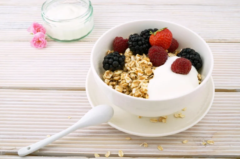 A white bowl of yogurt with granola, blackberries, raspberries, and a strawberry sits on a saucer with a spoon—perfect for National Nutrition Month inspiration—beside a small jar of yogurt and pink flowers on a wooden surface.