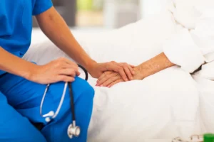 A healthcare worker in blue scrubs holds the hand of an elderly patient lying in bed, showing support—an example of compassion often seen when to consider assisted living. A stethoscope is visible in the worker’s other hand.