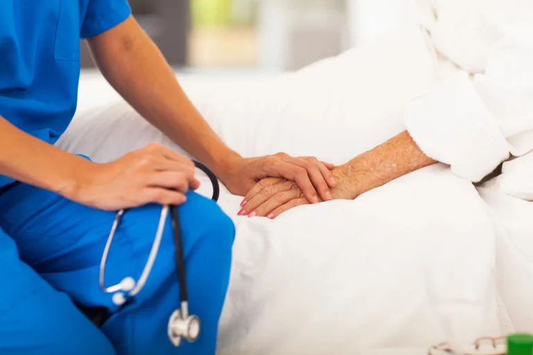 A healthcare worker in blue scrubs holds the hand of an elderly patient lying in bed, showing support—an example of compassion often seen when to consider assisted living. A stethoscope is visible in the worker’s other hand.