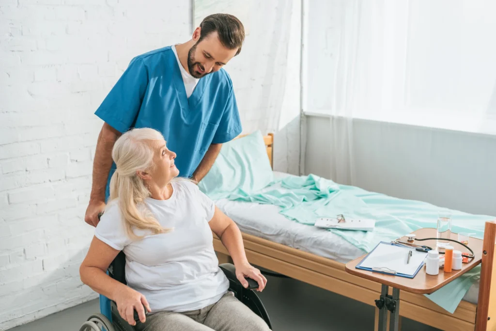 A nurse in blue scrubs talks to a woman in a wheelchair next to a hospital bed, with a clipboard and medicine bottles on a table nearby—discussing signs of assisted living and when to consider assisted living options.