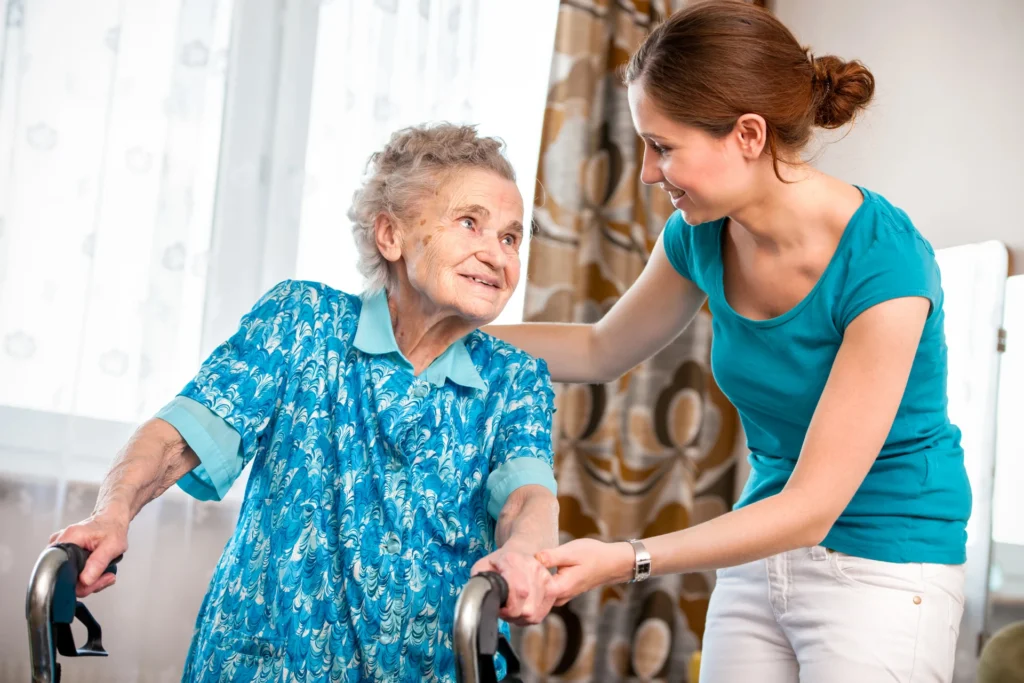 In a well-lit East Bay assisted living home, an elderly woman using a walker is helped by a younger woman in a turquoise shirt, both smiling warmly.