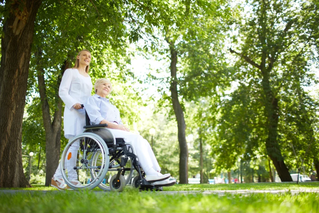 A younger woman pushes an older woman in a wheelchair through a sunny East Bay park with green trees and grass, highlighting the warmth of assisted living.