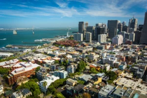Aerial view of East Bay cityscape featuring tall downtown buildings, a waterfront, and a suspension bridge under a partly cloudy sky—an inviting vista for those considering assisted living in the area.