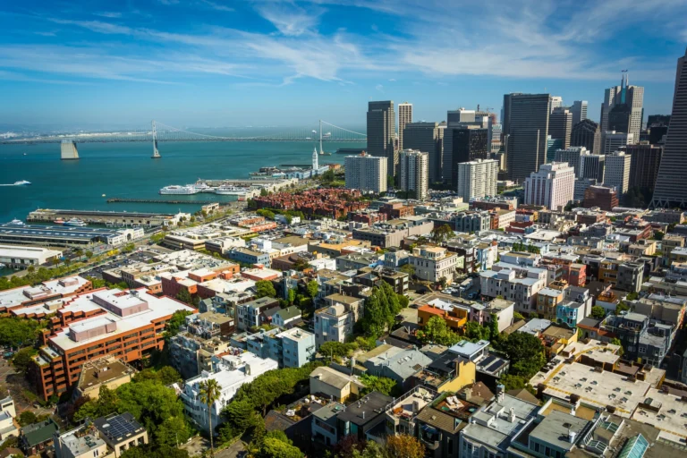 Aerial view of East Bay cityscape featuring tall downtown buildings, a waterfront, and a suspension bridge under a partly cloudy sky—an inviting vista for those considering assisted living in the area.