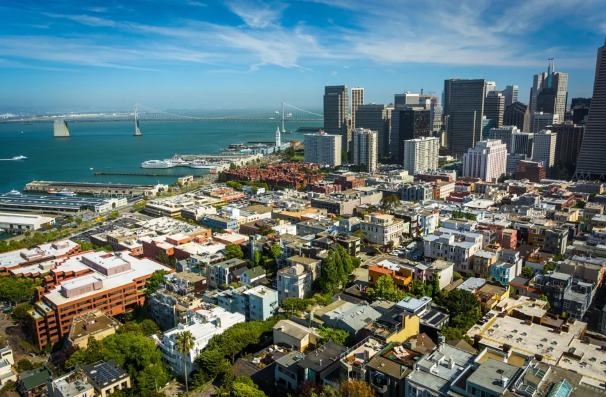 Aerial view of East Bay cityscape featuring tall downtown buildings, a waterfront, and a suspension bridge under a partly cloudy sky—an inviting vista for those considering assisted living in the area.