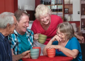 Four older adults sit around a table, laughing and holding mugs, in a cozy indoor setting with shelves and a red wall—enjoying the laughter benefits that contribute to their wellbeing.