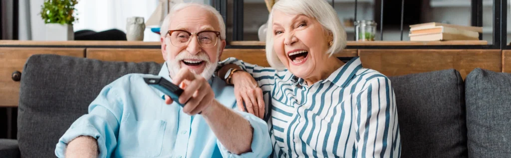 Two smiling older adults sit on a sofa, one holding a TV remote and pointing toward the camera, both appearing to enjoy watching television together while sharing the laughter benefits that boost their mood and health.