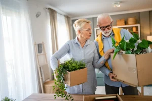 Two older adults, a woman and a man, stand indoors holding cardboard boxes with green potted plants, appearing to be moving or downsizing in a well-lit living room.