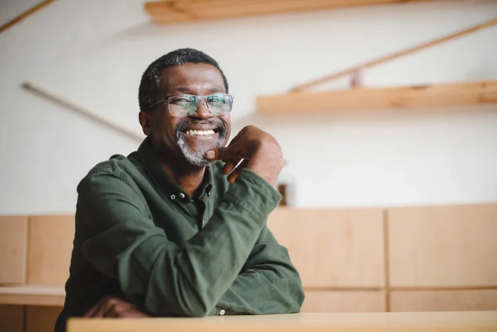 Man with short hair, glasses, and a gray beard sits at a table, smiling, wearing a green shirt — the picture of independence and contentment in senior living — with light wooden shelves and a white wall in the background.