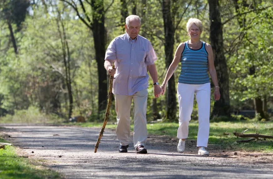 An older man and woman walk hand in hand on a paved path through a wooded area, embodying the spirit of independence often enjoyed in senior living. The man uses a walking stick, and both are smiling and dressed in light, casual clothing.