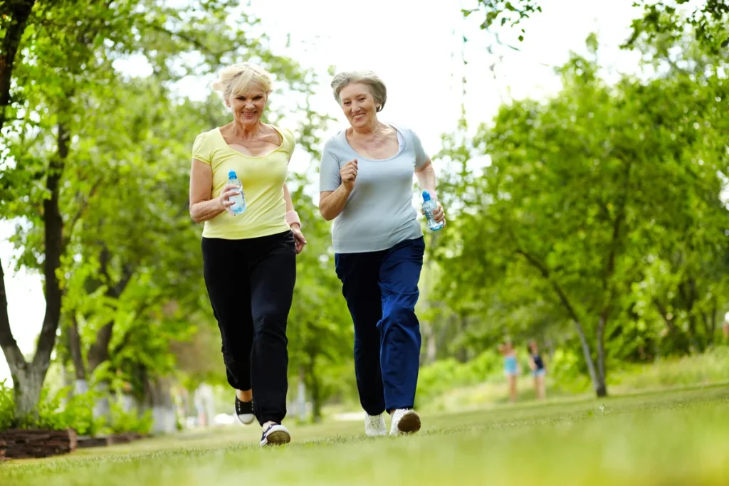 Two older women wearing athletic clothes jog together in a green park, each holding a water bottle, with trees and grass surrounding them on a sunny day. This scene highlights safe exercise for seniors, showing how outdoor fitness for seniors can be both enjoyable and healthy.