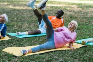 Group of seniors exercising on mats outdoors in July, lying on their sides and lifting one leg, with trees and grass in the background for a refreshing outdoor fitness session.