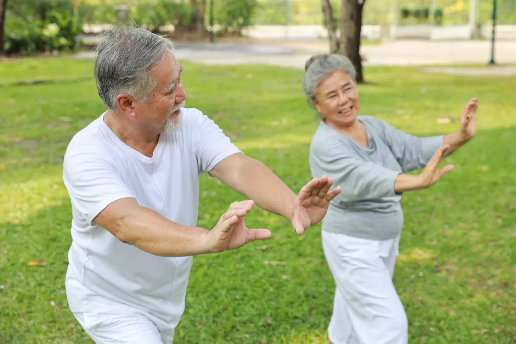 Two older adults practicing tai chi outdoors on grass, both smiling and wearing comfortable clothing, demonstrate safe outdoor exercise as part of outdoor fitness for seniors.