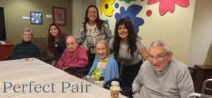 Four elderly adults and three younger adults pose and smile around a table in a brightly decorated room with abstract wall art, capturing the spirit of Bridging Generations. The words “Perfect Pair” appear in the lower left corner.