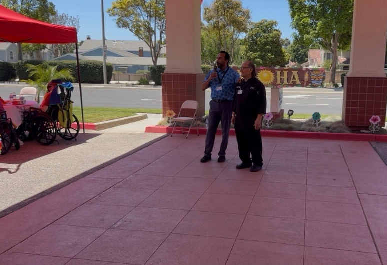 Two men stand under a covered outdoor area at AlmaVia of Camarillo one holding a microphone perhaps addressing Victor Camachos retirement A table with a pink tablecloth and wheelchairs are on the left while the background features trees a sign and a street