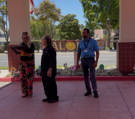 Three adults stand outside near a building entrance one woman holds a framed document while the two men including Victor Camacho of AlmaVia of Camarillo stand nearby Trees and a decorated sign are visible in the background suggesting the celebration of a retirement milestone