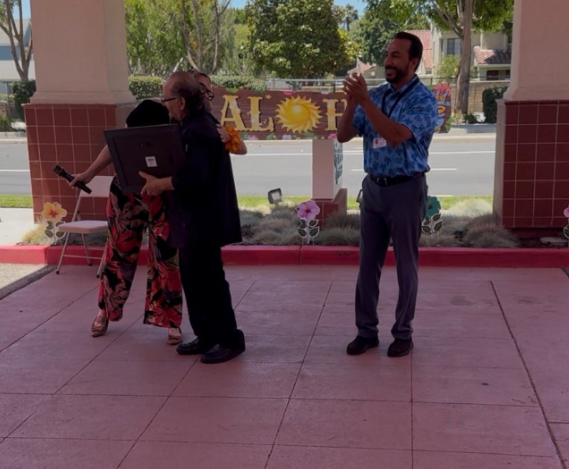 Three people stand under a covered outdoor area at AlmaVia of Camarillo one holds a large framed item another holds a microphone and a third person is clapping The word ALOHA is displayed in the background as Victor Camacho is honored during his retirement celebration