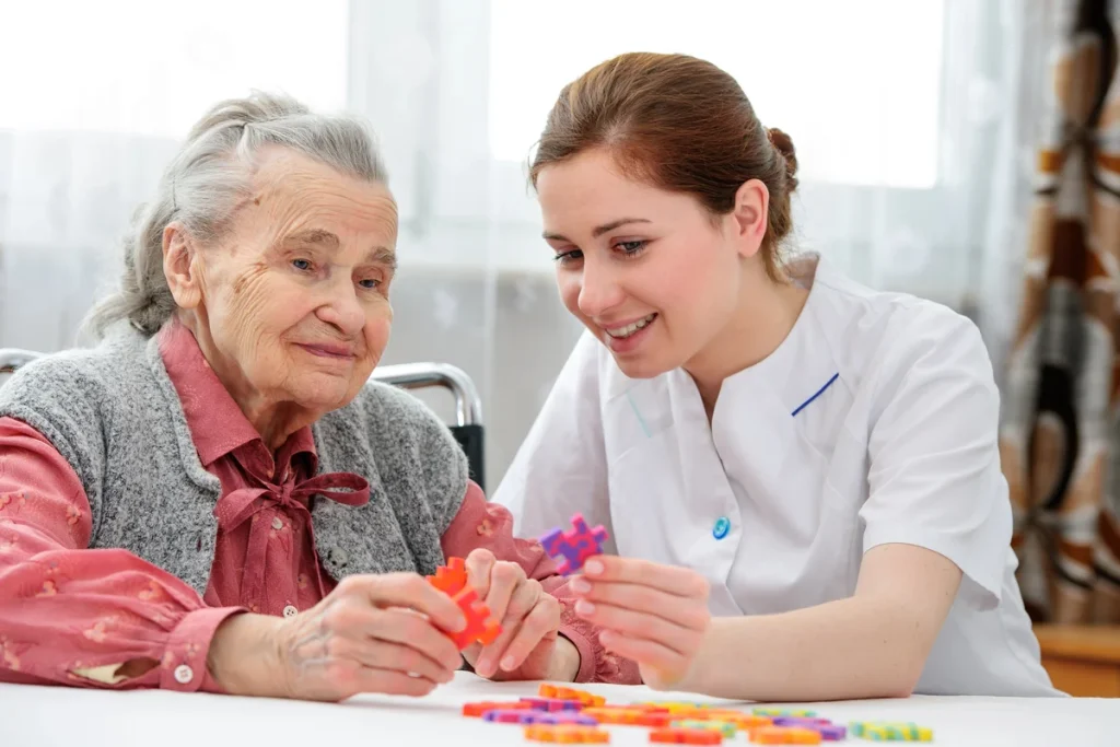 An elderly woman and a caregiver work together to assemble a colorful puzzle at a table in a bright room, enjoying meaningful socialization for older adults that supports healthy aging.