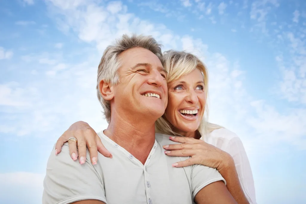 Smiling older couple outdoors, the woman hugs the man from behind as they both look into the distance under a blue sky with scattered clouds. Their joyful connection is a beautiful reflection of socialization for older adults—a vital component of healthy aging.