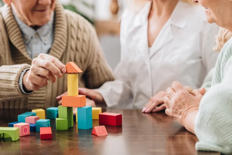 Two older adults and a caregiver sit at a table, building a colorful structure with wooden blocks—an engaging activity that supports brain health after 65.