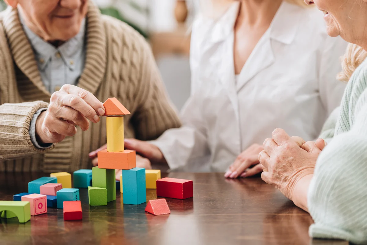 Two older adults and a caregiver sit at a table, building a colorful structure with wooden blocks—an engaging activity that supports brain health after 65.