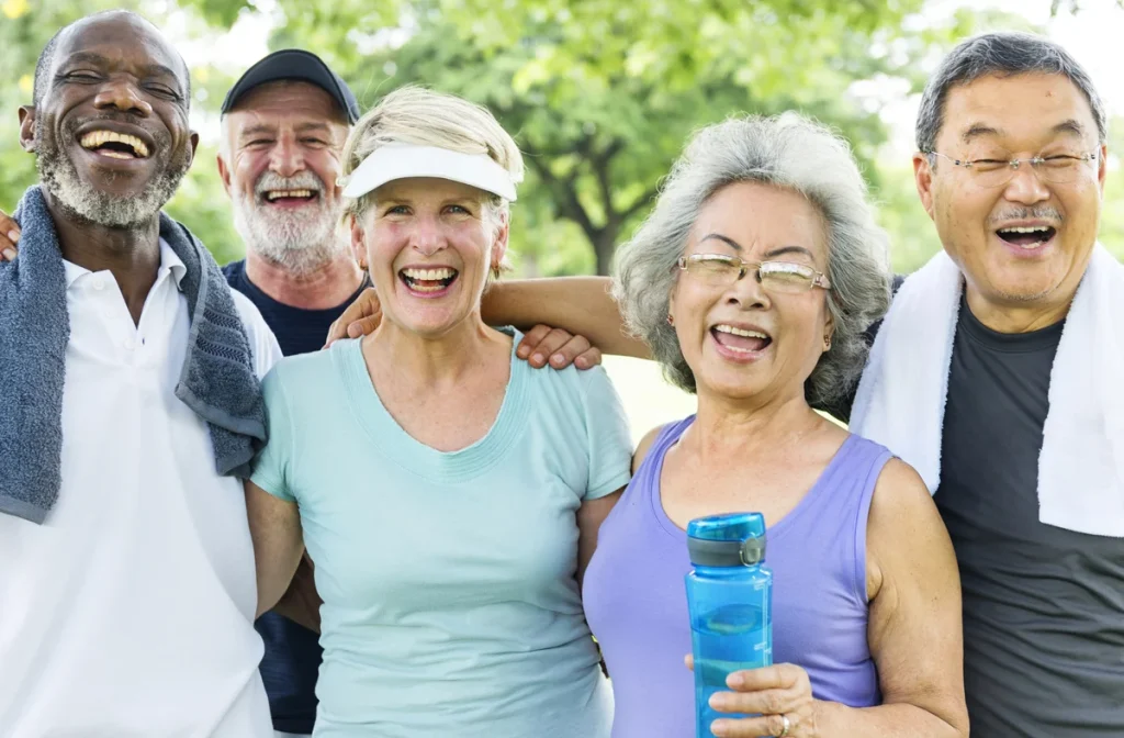 Five older adults stand outdoors in casual workout clothes, smiling and laughing together, with trees and greenery in the background, enjoying an active lifestyle as part of a vibrant nonprofit senior living community.