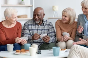 Four older adults sit together at a table in a cheerful senior living community, smiling and playing cards. Cups, snacks, and a remote control are on the table in front of them, capturing the warm atmosphere of nonprofit senior housing.