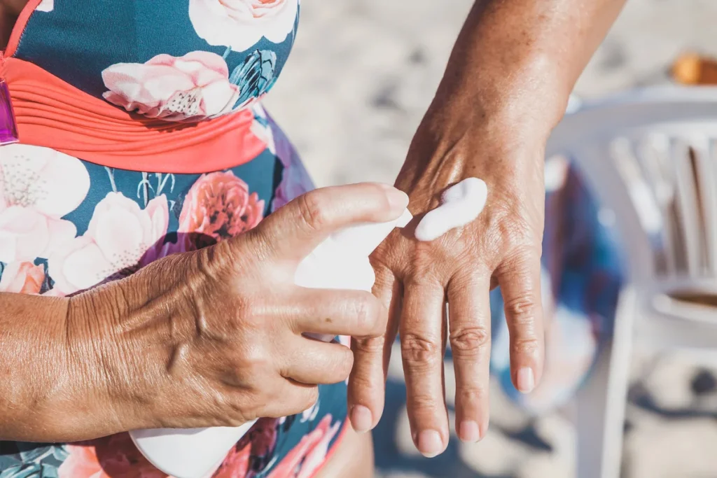 A person in a floral swimsuit applies sunscreen lotion to the back of their hand while sitting outdoors, practicing sun safety, with a plastic chair visible in the background.