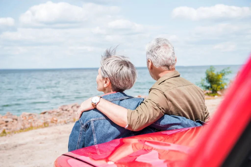 Two older adults sit side by side, facing a large body of water, with one person’s arm around the other beside a red car, under a partly cloudy sky. They enjoy the peaceful moment while practicing sun safety, ensuring they’re protected even on days when sun myths might suggest clouds provide enough shade.