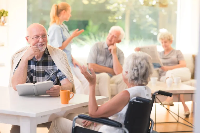 A man sits at a table reading a book and smiling, talking to a woman in a wheelchair. Other seniors are in the background in a bright common room, capturing a typical day in assisted living.