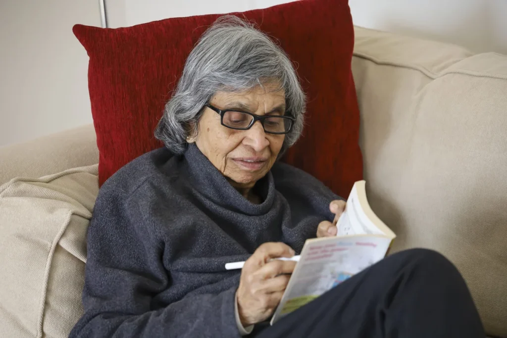 An older woman with gray hair and glasses sits on a couch, writing in a book with a pen. A red pillow is behind her back, capturing a peaceful moment from her typical day in assisted living.
