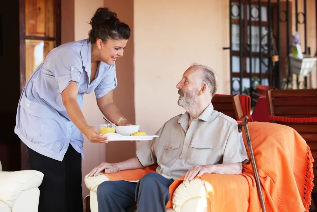 A woman hands a tray with food and drinks to an older man seated in an armchair with an orange blanket, in a home setting, capturing a typical day in assisted living and senior care.