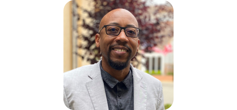 Cleitus Jones, Executive Director at AlmaVia of San Francisco, is a smiling man with a goatee and glasses, wearing a light-colored blazer over a dark collared shirt.