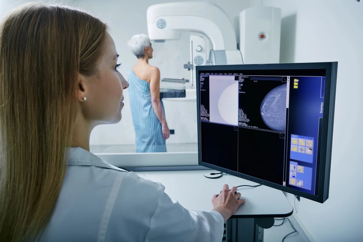 A technician reviews a mammogram on a computer monitor for cancer prevention while a patient stands by the imaging machine.