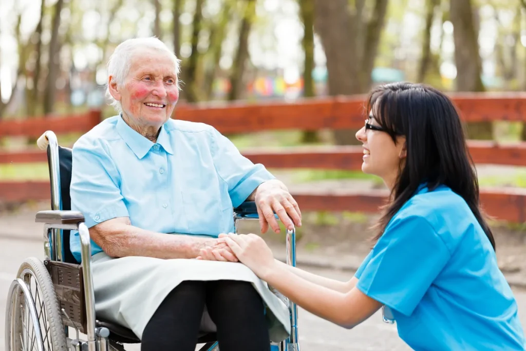 A caregiver in blue scrubs holds the hands of an elderly woman in a wheelchair during her hospital stay.