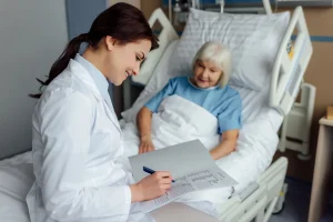 A doctor in a white coat writes on a medical chart while discussing post-hospital care with an elderly patient in a hospital bed.