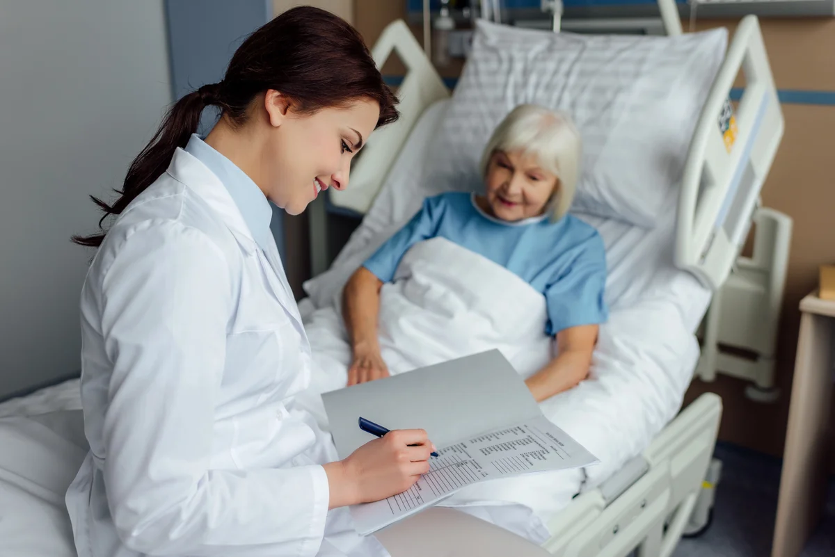 A doctor in a white coat writes on a medical chart while discussing post-hospital care with an elderly patient in a hospital bed.