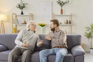 An older man and a younger man sit on a sofa, talking about senior care planning.